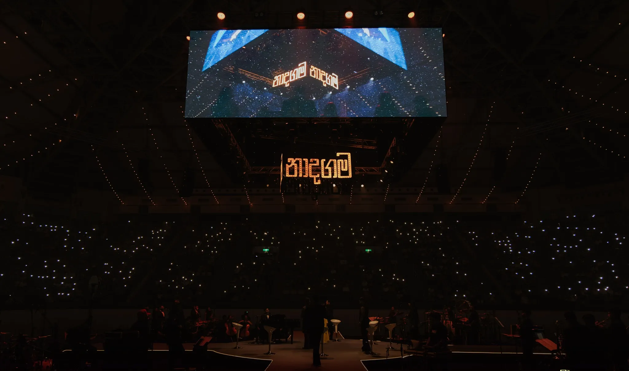 An arena lit by phone lights beneath the glowing Naadha Gama sign